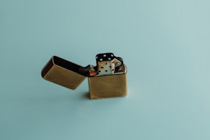 A brass-colored metal lighter with its lid open against a light blue background. The lighter has a slightly worn and aged appearance.