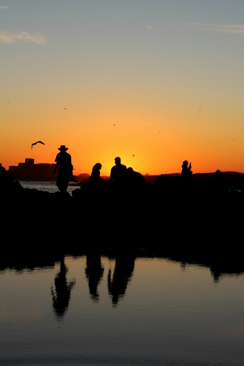 silhouette of people standing on rock formation near body of water during sunset