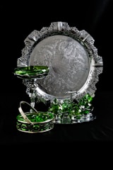 An ornate silver tray with intricate patterns and a decorative border is set against a dark background. In front of the tray, there are several green glass pieces, including a pedestal dish, stacked cups, and a basket with a handle. The reflective surfaces create a contrast between the shiny metal and the translucent green glass.