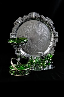 An ornate silver tray with intricate patterns and a decorative border is set against a dark background. In front of the tray, there are several green glass pieces, including a pedestal dish, stacked cups, and a basket with a handle. The reflective surfaces create a contrast between the shiny metal and the translucent green glass.