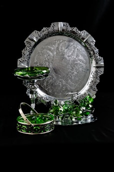 An ornate silver tray with intricate patterns and a decorative border is set against a dark background. In front of the tray, there are several green glass pieces, including a pedestal dish, stacked cups, and a basket with a handle. The reflective surfaces create a contrast between the shiny metal and the translucent green glass.