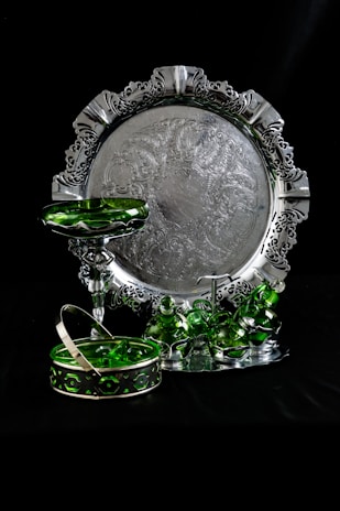 An ornate silver tray with intricate patterns and a decorative border is set against a dark background. In front of the tray, there are several green glass pieces, including a pedestal dish, stacked cups, and a basket with a handle. The reflective surfaces create a contrast between the shiny metal and the translucent green glass.