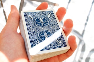 A hand holding a deck of playing cards, with the back design featuring intricate blue and white artwork including angles and patterns. The lighting is bright, creating a soft shadow and highlighting the details.