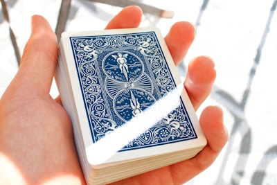 A hand holding a deck of playing cards, with the back design featuring intricate blue and white artwork including angles and patterns. The lighting is bright, creating a soft shadow and highlighting the details.