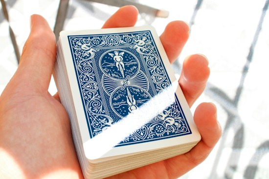 A hand holding a deck of playing cards, with the back design featuring intricate blue and white artwork including angles and patterns. The lighting is bright, creating a soft shadow and highlighting the details.