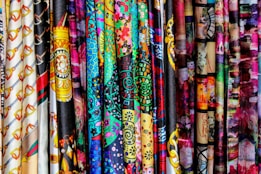 A vibrant assortment of fabrics is displayed, each featuring unique and intricate patterns. The patterns include various shapes, designs, and colors, ranging from geometric to floral motifs, set against both dark and bright backgrounds.