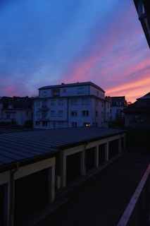 A residential neighborhood at dusk featuring a prominent building against a dramatic sky. The sunset casts vibrant shades of blue, pink, and orange across the horizon. The scene includes rows of garage rooftops, with some dimly lit windows visible in the building.
