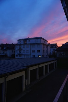 A residential neighborhood at dusk featuring a prominent building against a dramatic sky. The sunset casts vibrant shades of blue, pink, and orange across the horizon. The scene includes rows of garage rooftops, with some dimly lit windows visible in the building.