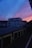 A residential neighborhood at dusk featuring a prominent building against a dramatic sky. The sunset casts vibrant shades of blue, pink, and orange across the horizon. The scene includes rows of garage rooftops, with some dimly lit windows visible in the building.