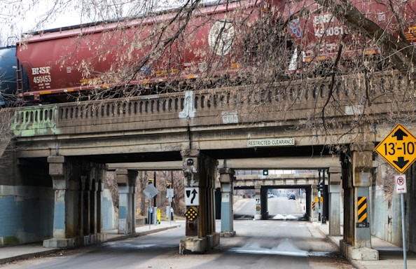 A freight train with red and blue cars moves along a concrete bridge that spans over a narrow road. The bridge features signs indicating restricted clearance and a height warning of 13 feet and 10 inches. Leafless tree branches hang above and around the bridge, adding to the urban environment, with traffic signs and road markings visible below.
