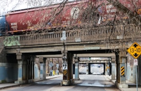 A freight train with red and blue cars moves along a concrete bridge that spans over a narrow road. The bridge features signs indicating restricted clearance and a height warning of 13 feet and 10 inches. Leafless tree branches hang above and around the bridge, adding to the urban environment, with traffic signs and road markings visible below.