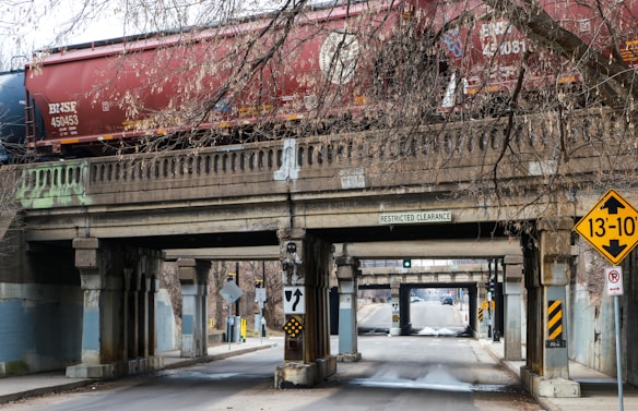 A freight train with red and blue cars moves along a concrete bridge that spans over a narrow road. The bridge features signs indicating restricted clearance and a height warning of 13 feet and 10 inches. Leafless tree branches hang above and around the bridge, adding to the urban environment, with traffic signs and road markings visible below.
