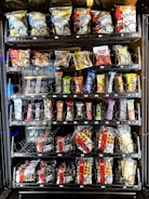 A vending machine offering a variety of snacks, including chips, candies, and waffles. The shelves are neatly organized with multiple rows displaying different brands and flavors. Snack packets are secured behind spiraled wire mechanisms designed to dispense items upon selection.