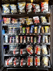 A vending machine offering a variety of snacks, including chips, candies, and waffles. The shelves are neatly organized with multiple rows displaying different brands and flavors. Snack packets are secured behind spiraled wire mechanisms designed to dispense items upon selection.