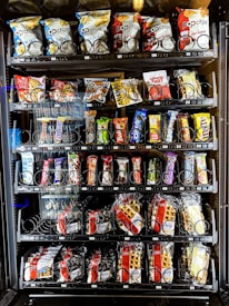 A vending machine offering a variety of snacks, including chips, candies, and waffles. The shelves are neatly organized with multiple rows displaying different brands and flavors. Snack packets are secured behind spiraled wire mechanisms designed to dispense items upon selection.