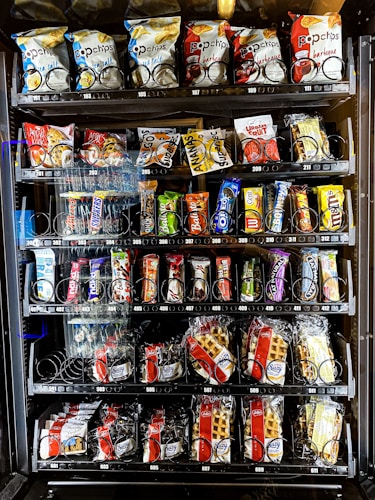 A vending machine offering a variety of snacks, including chips, candies, and waffles. The shelves are neatly organized with multiple rows displaying different brands and flavors. Snack packets are secured behind spiraled wire mechanisms designed to dispense items upon selection.