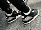 A pair of black and white athletic shoes with a distinctive pattern on the heel and the Nike Air logo displayed. The shoes are worn by someone clothing in dark pants and black socks, seated on a carpeted floor.