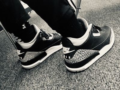 A pair of black and white athletic shoes with a distinctive pattern on the heel and the Nike Air logo displayed. The shoes are worn by someone clothing in dark pants and black socks, seated on a carpeted floor.