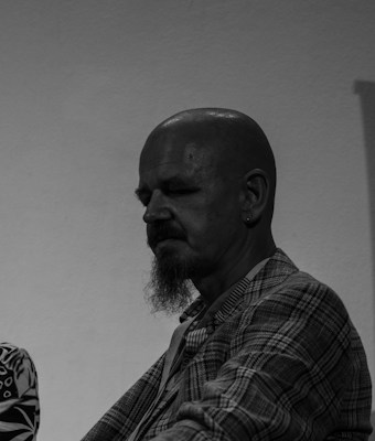 A grayscale portrait of a bald man with a mustache and goatee, wearing a plaid jacket. He has an earring and is gazing downward, creating a contemplative expression. The background is plain and minimalistic.