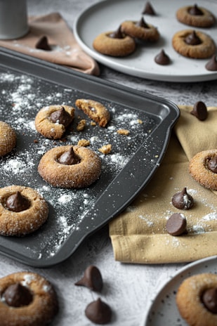 Freshly baked cookies topped with chocolate kisses are placed on a dark baking tray, dusted with powdered sugar. Surrounding the tray are additional cookies and chocolate kisses on light-colored plates and cloths, some scattered crumbs, and a white cup in the background.