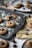 Freshly baked cookies topped with chocolate kisses are placed on a dark baking tray, dusted with powdered sugar. Surrounding the tray are additional cookies and chocolate kisses on light-colored plates and cloths, some scattered crumbs, and a white cup in the background.