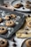 Freshly baked cookies topped with chocolate kisses are placed on a dark baking tray, dusted with powdered sugar. Surrounding the tray are additional cookies and chocolate kisses on light-colored plates and cloths, some scattered crumbs, and a white cup in the background.