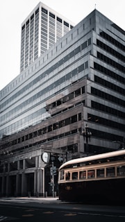 A modern urban scene with a large, multi-story glass and steel office building. In the foreground, there is a vintage-style streetcar on tracks, hinting at a blend of the old and new in the cityscape. Street signs and a lamp post are also visible.