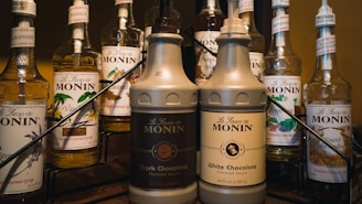 A variety of Monin syrup bottles are displayed, featuring different flavors including lavender, vanilla, caramel, peppermint, and toasted marshmallow. Two large pumps labeled as dark chocolate and white chocolate flavored sauces are prominently in the foreground. The bottles are arranged on a metal rack and have colorful labels indicative of their respective flavors.