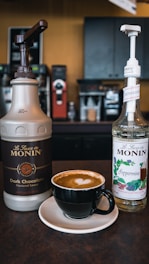A cup of coffee with a heart latte art on top sits on a saucer, flanked by two bottles of Monin syrup: one for dark chocolate flavored sauce and the other for peppermint syrup. They are placed on a countertop in a coffee shop setting with an espresso machine visible in the background.