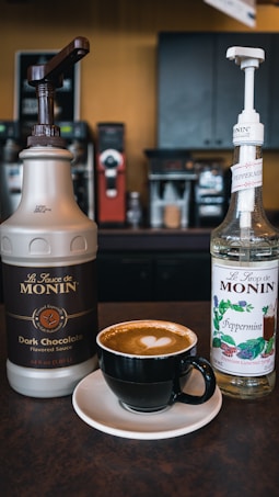 A cup of coffee with a heart latte art on top sits on a saucer, flanked by two bottles of Monin syrup: one for dark chocolate flavored sauce and the other for peppermint syrup. They are placed on a countertop in a coffee shop setting with an espresso machine visible in the background.