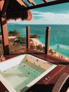 An outdoor hot tub is set on a wooden deck with a thatched roof offering a breathtaking view of the ocean. The clear blue water stretches to the horizon, with small boats visible in the distance. A white sandy beach lined with lush green trees is below, creating a tropical paradise atmosphere.
