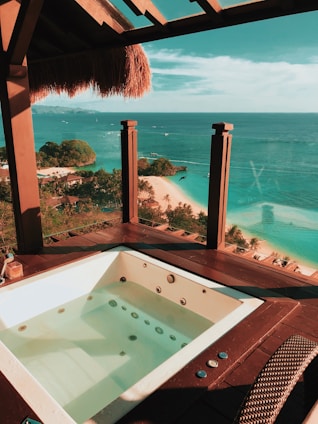 An outdoor hot tub is set on a wooden deck with a thatched roof offering a breathtaking view of the ocean. The clear blue water stretches to the horizon, with small boats visible in the distance. A white sandy beach lined with lush green trees is below, creating a tropical paradise atmosphere.