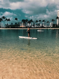 A serene coastal scene featuring an individual paddleboarding on calm, shallow waters. In the background, several people engage in water activities, set against a backdrop of palm trees and a cityscape with tall buildings. The sky is partly cloudy, adding to the tranquil atmosphere.