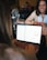 A person is working on a laptop with a calendar app open. Another person, holding a phone and a notebook, is sitting across the table, smiling. There are drinks on the table, a transparent water bottle visible, and several people are in the background.