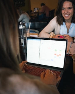 A person is working on a laptop with a calendar app open. Another person, holding a phone and a notebook, is sitting across the table, smiling. There are drinks on the table, a transparent water bottle visible, and several people are in the background.