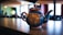 A colorful, intricately patterned teapot sits on a dark countertop in a warmly lit interior space. The teapot features a vivid blue base with red, yellow, and white floral designs. The background is softly blurred, showing hints of a cozy cafe or home setting with muted green and brown tones and large windows letting in light.