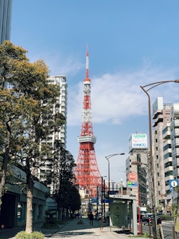 A tall red and white broadcasting tower rises amidst a cityscape, surrounded by modern buildings and leafy trees. The sky is mostly clear with a few clouds. People walk along the sidewalk, and a taxi stand is visible.