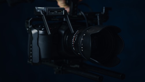 A professional video camera with a large lens is shown, equipped with additional gear for stability and functionality in low-light conditions. The camera body is sleek and predominantly black, with various dials and buttons visible for manual control.
