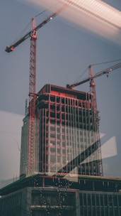 A tall construction site featuring a high-rise building with two large cranes. The building structure shows exposed steel beams and partially completed floors, while the cranes tower above, indicating ongoing development. The photo is taken through a window with visible reflections.
