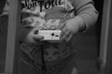 A small child holds a digital camera. The child is wearing a shirt with cartoon graphics and heart patterns. The sleeves are long, and part of the shirt reads 'on Tour' with a visible cartoon character. The scene is captured in grayscale, focusing on the child's hands and torso.