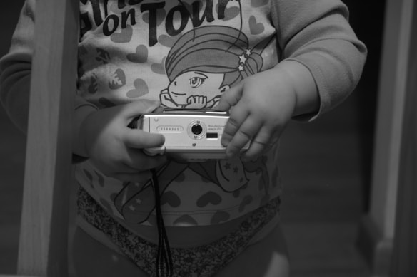 A small child holds a digital camera. The child is wearing a shirt with cartoon graphics and heart patterns. The sleeves are long, and part of the shirt reads 'on Tour' with a visible cartoon character. The scene is captured in grayscale, focusing on the child's hands and torso.
