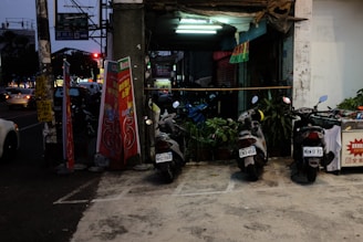 Three scooters are parked next to each other in a dimly lit parking area in front of a shop. Several potted plants are placed behind the scooters. The shop's entrance is illuminated by a fluorescent light, and two red and white advertisement boards are placed on the left side. Signs written in a non-English language are visible. In the background on the street, there are cars and a traffic light.