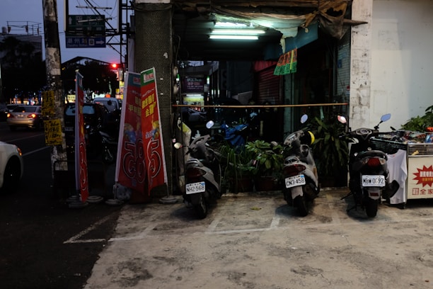 Three scooters are parked next to each other in a dimly lit parking area in front of a shop. Several potted plants are placed behind the scooters. The shop's entrance is illuminated by a fluorescent light, and two red and white advertisement boards are placed on the left side. Signs written in a non-English language are visible. In the background on the street, there are cars and a traffic light.