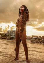 A person stands confidently by the edge of the water, wearing reflective sunglasses and a metallic bikini with a sheer cover-up. The background features a beach scene with buildings, palm trees, and thatched umbrellas, under a golden, cloudy sky.