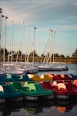 A row of colorful paddle boats in red, yellow, and green are docked alongside a body of water. In the background, several sailboats with tall masts are lined up, with trees and a clear sky completing the calm setting.