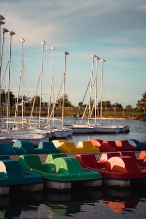 A row of colorful paddle boats in red, yellow, and green are docked alongside a body of water. In the background, several sailboats with tall masts are lined up, with trees and a clear sky completing the calm setting.
