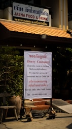 A restaurant sign with the name 'JAENA FOOD & DRINK' indicates a temporary closure for dine-in services due to COVID-19. The notice communicates the availability of takeout services only. The sign is positioned near potted plants and seating outside the restaurant.