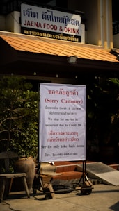 A restaurant sign with the name 'JAENA FOOD & DRINK' indicates a temporary closure for dine-in services due to COVID-19. The notice communicates the availability of takeout services only. The sign is positioned near potted plants and seating outside the restaurant.