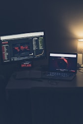 A dimly lit room with a desk featuring two screens displaying global COVID-19 statistics. A large monitor shows a world map with colored markers indicating various locations, alongside numerical data and graphs. A laptop is positioned nearby, also displaying a map with data, and an open notebook sits on the desk. A small lamp with a subtle glow provides additional lighting.