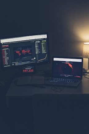 A dimly lit room with a desk featuring two screens displaying global COVID-19 statistics. A large monitor shows a world map with colored markers indicating various locations, alongside numerical data and graphs. A laptop is positioned nearby, also displaying a map with data, and an open notebook sits on the desk. A small lamp with a subtle glow provides additional lighting.